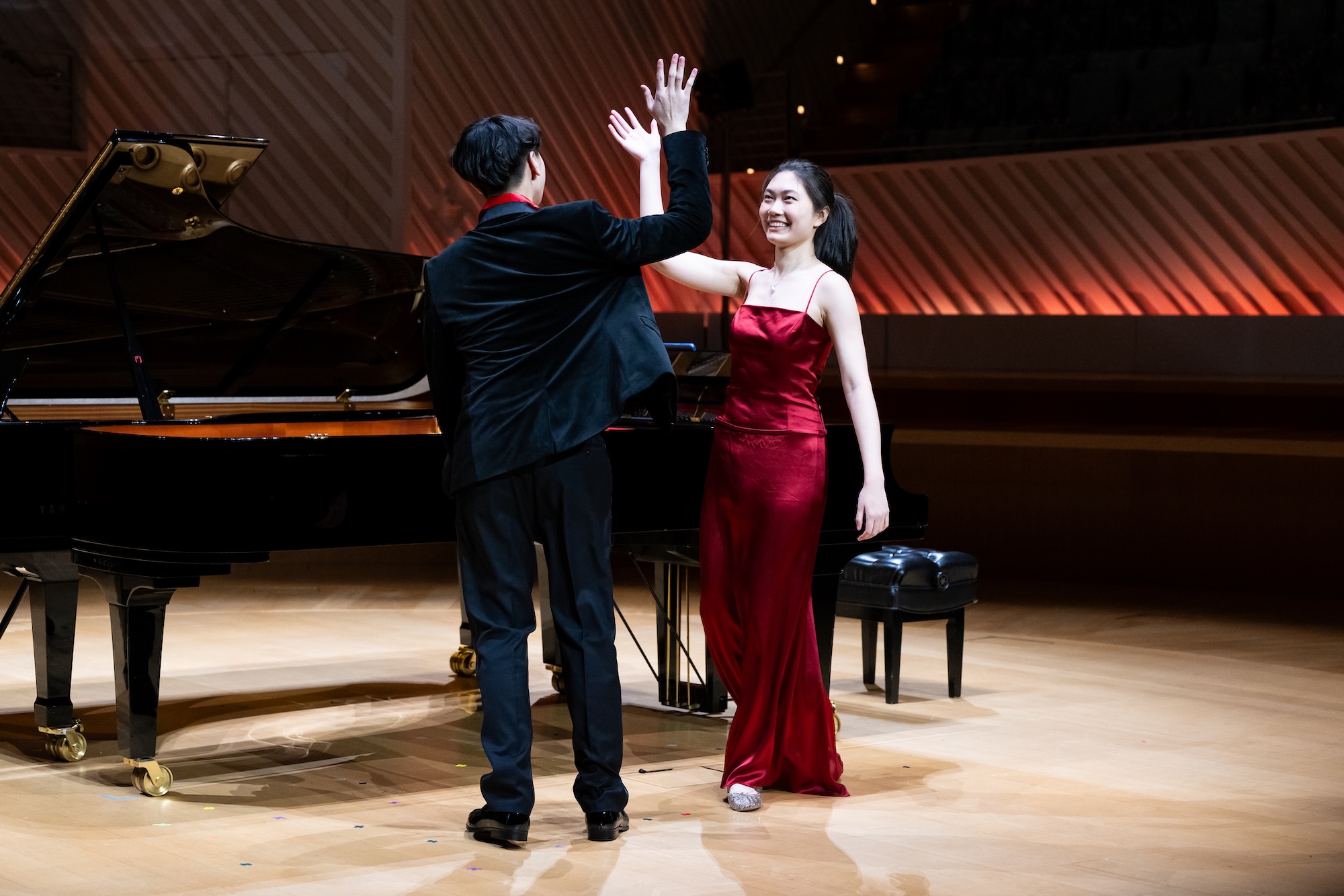 Two young musicians raise their hands beside grand pianos on a concert hall stage.