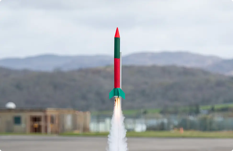 Red and green model rocket launching with a bright exhaust plume against a blurred outdoor background.