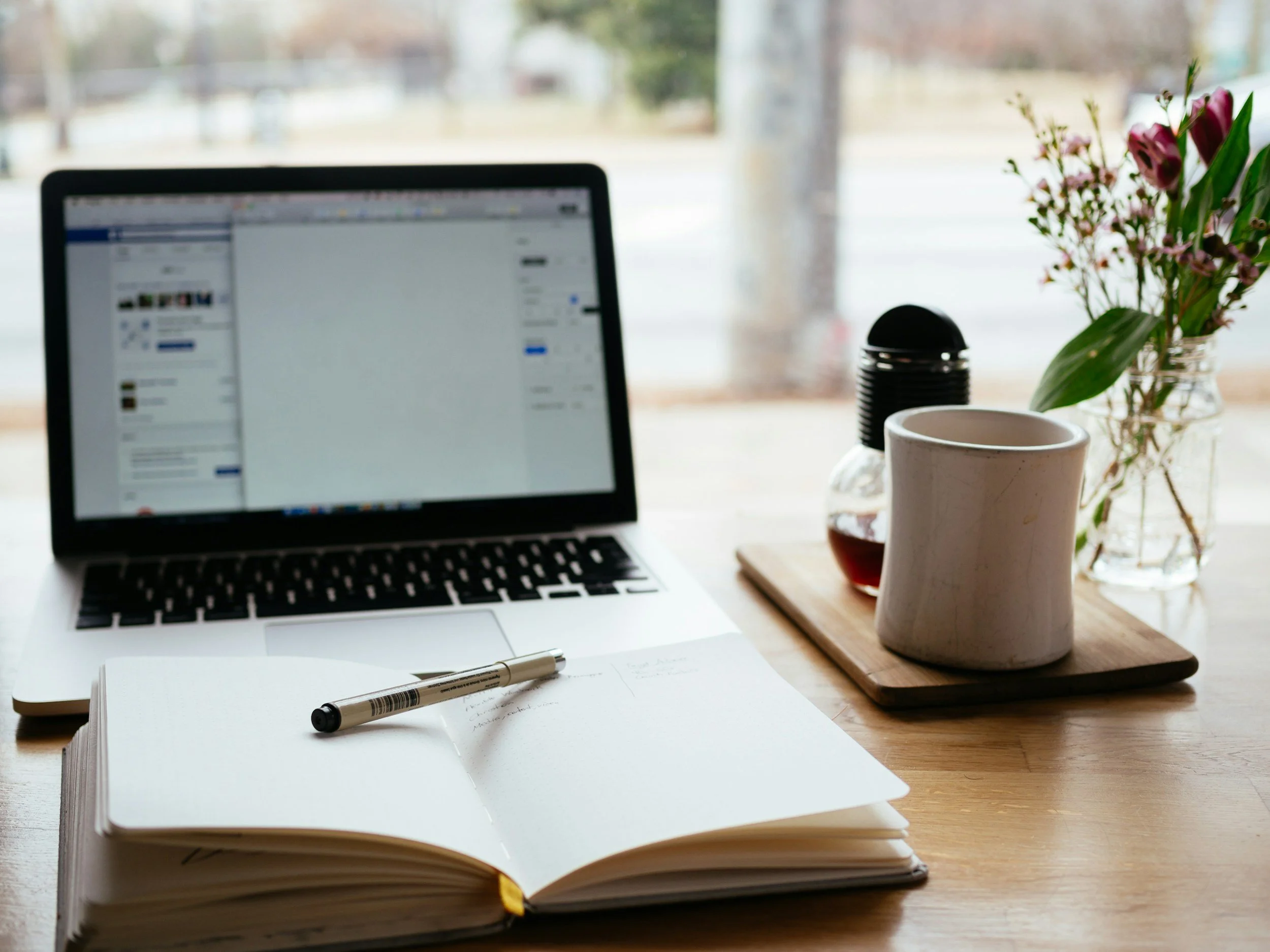 Open notebook and laptop on a desk beside a mug and flowers