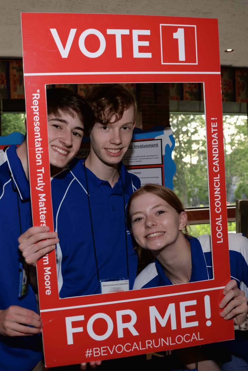 Three students posing with a red Vote 1 campaign frame at a Community Projects event.