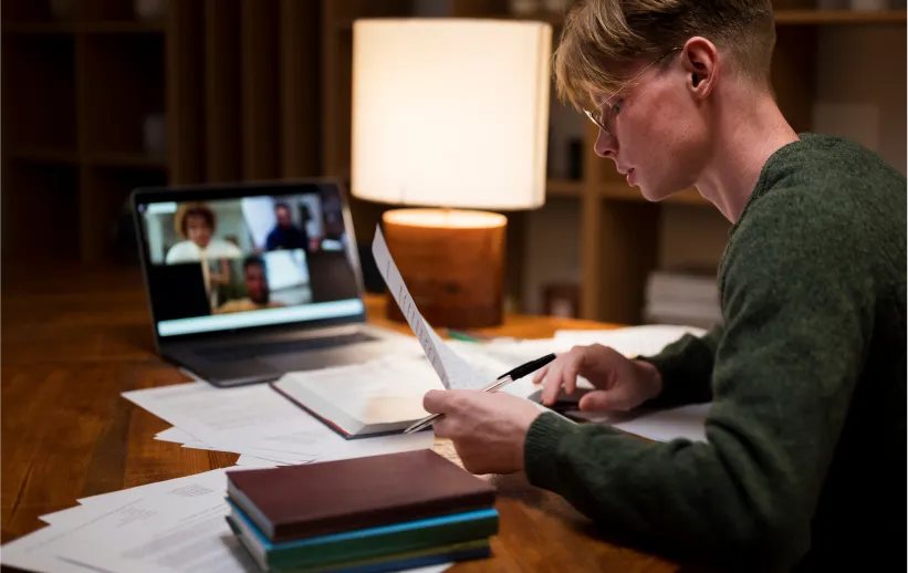 Young person reviewing papers with a pen at a desk beside books and a laptop showing a video call.