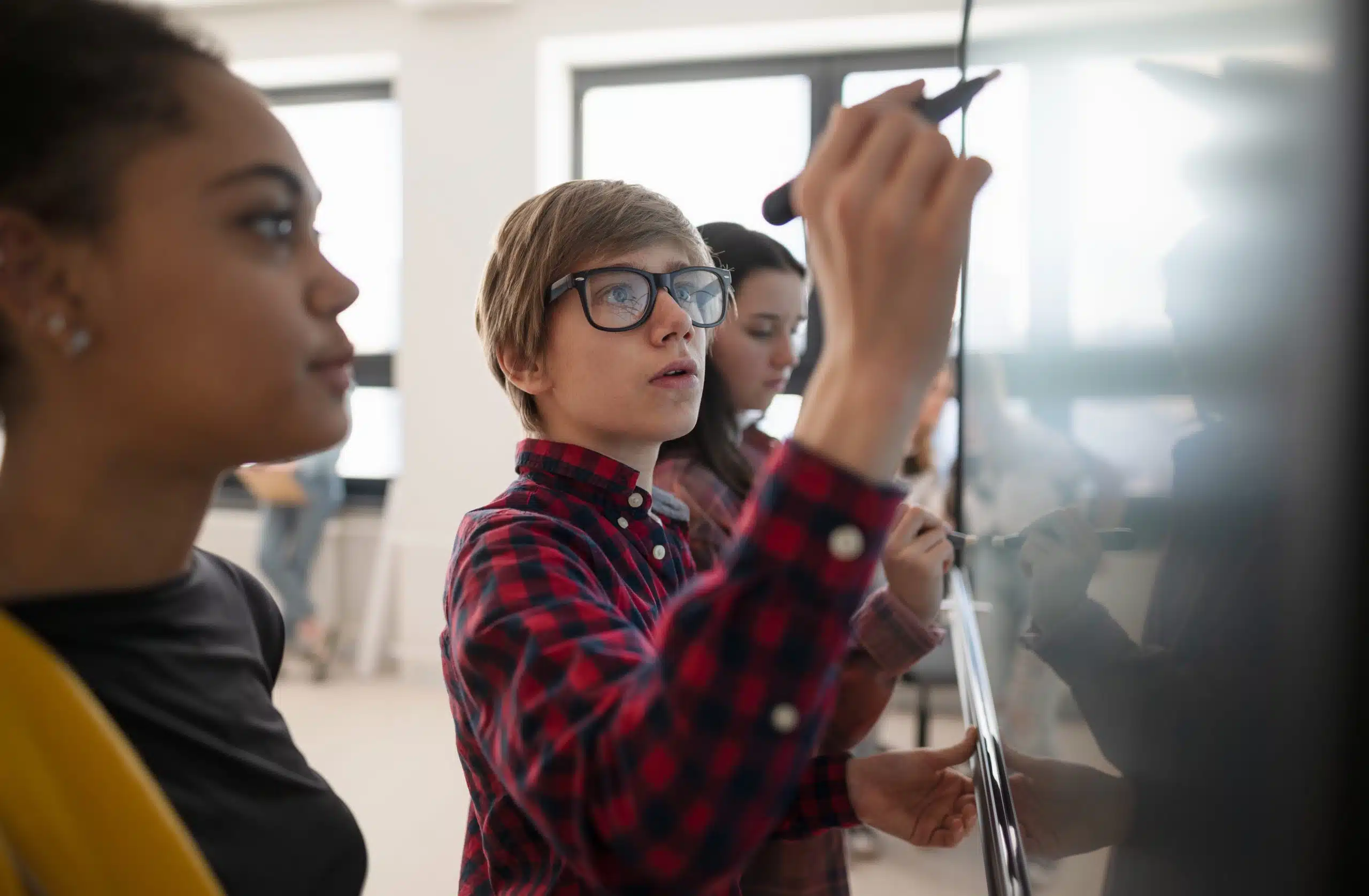 Student wearing glasses writing on a classroom board while classmates watch